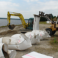 Befüllung mit lokal vorhandenem Sand der SoilTain Bags mit Hilfe eines Baggers während Radlader den vorkonfektionierten Sandsack aufhält