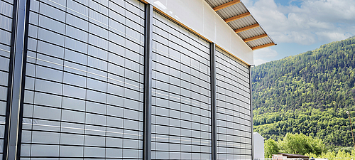 Tectura Faltfron 3 große Faltfronten an einer Lagerhalle. Im Hintergrund sind bepflanzte Berge und blauer Himmer zu erkennen.