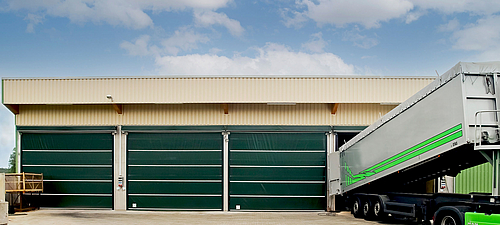 Lubratec Stabitore Drei geschlossene grüne Lubratec Stabitore an einer beigen Lagerhalle. Am rechten geöffneten Tor wird ein LKW entladen.