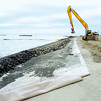 Ein Bagger verkleidet eingebaute Erosionsschutzmatten mithilfe von Schüttsteindeckwerk am Meeresufer